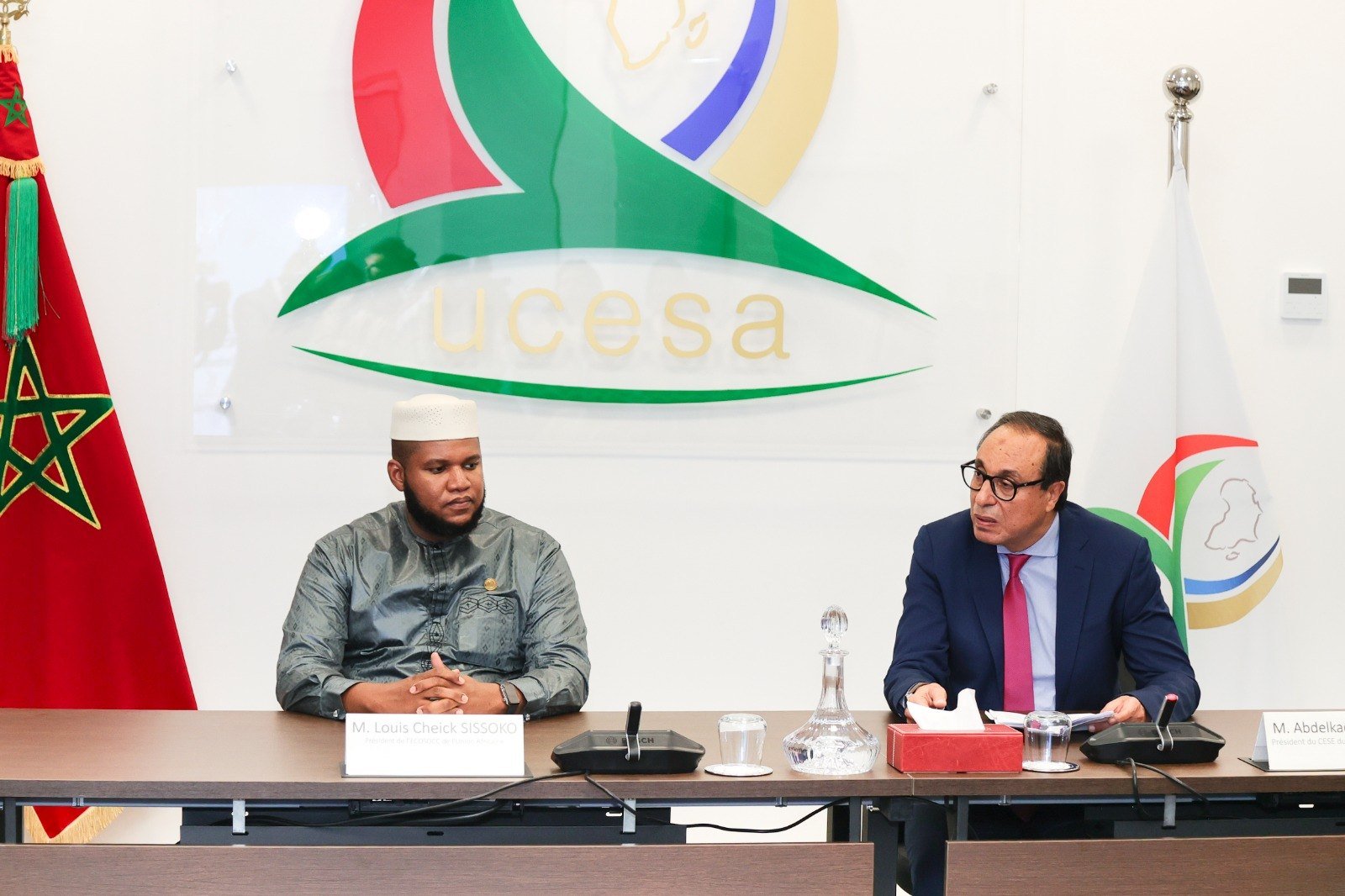 شخصان يجلسان على طاولة مؤتمر مع شعار UCESA والأعلام الوطنية في الخلفية