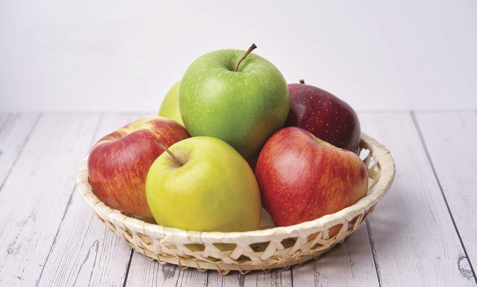 Colorful fresh apples in a woven basket on white wooden table