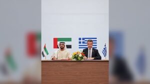 Two officials seated at desk signing agreement with UAE and Greek flags