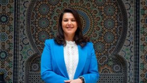 امرأة بملابس رسمية زرقاء وبيضاء أمام خلفية معمارية إسلامية مزخرفة