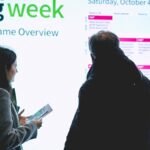 Dos asistentes de pie frente a pantalla del programa UEG Week, uno sostiene material impreso